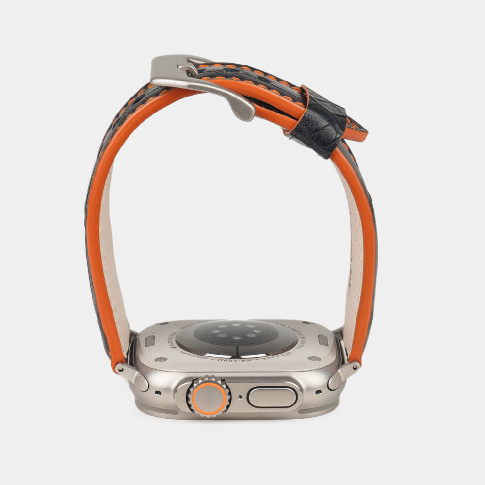 Apple Watch Ultra 49 mm in black alligator leather with orange stitching and edge, with titanium adapters and buckle | watch case on bottom to see the orange dyed edge