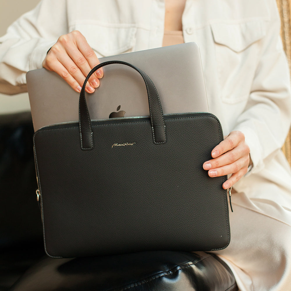 A person in a white shirt holds a black pebbled leather briefcase-style laptop bag with top handles, shown in a real indoor setting with a laptop behind it.