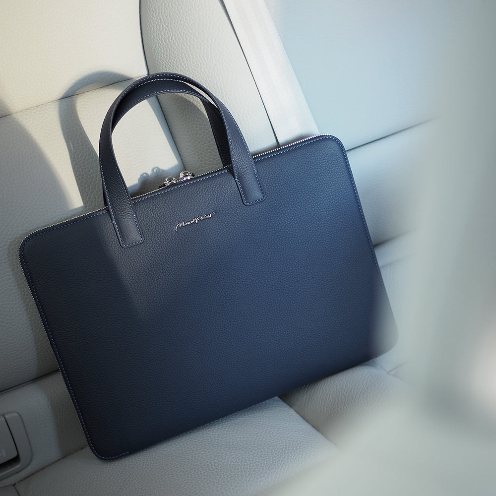 A navy blue pebbled leather briefcase with top handles resting on a light-colored leather car seat.