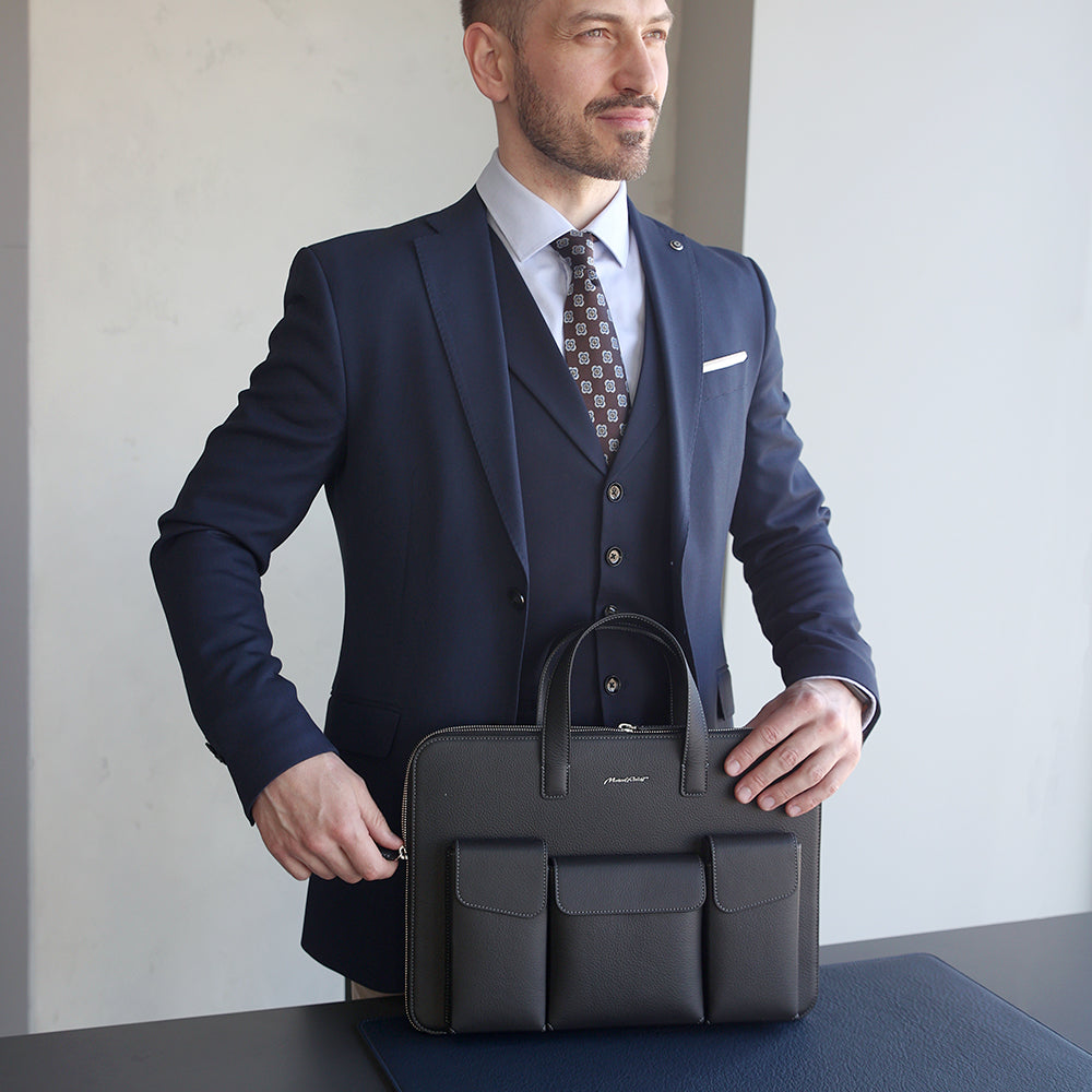 A man in a navy blue three-piece suit holds a black leather briefcase with three front pockets