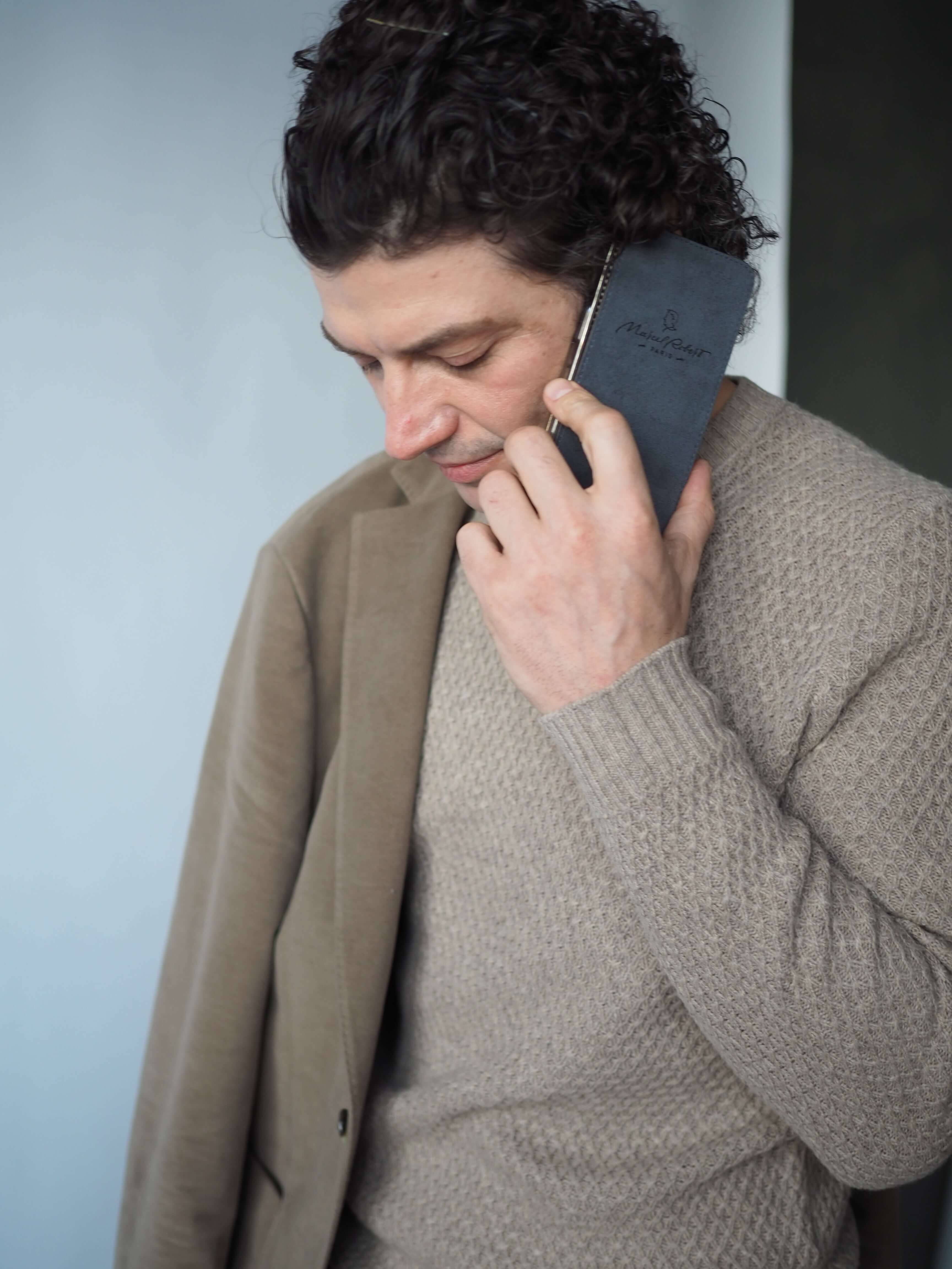 Man talking on a phone with a phone leather flip case featuring a brand logo, wearing a beige sweater and coat.