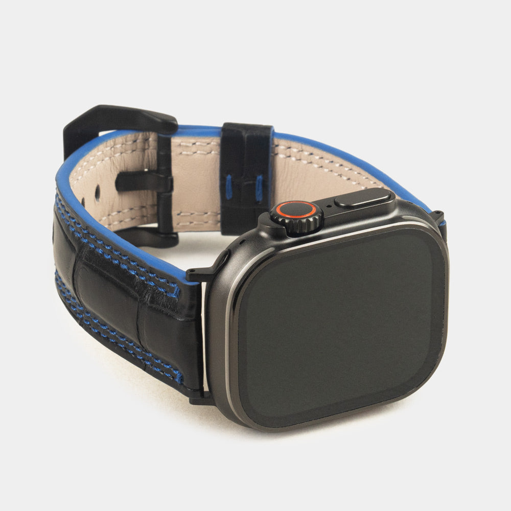 Apple Watch Ultra 49 mm in black alligator leather with blue stitching and edge, with black adapters and buckle | 3/4 front view showing the black watch case and the square scales of the leather