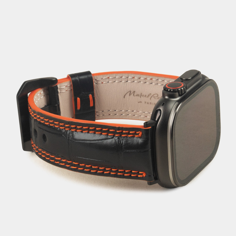 Apple Watch Ultra 49 mm in black alligator leather with orange stitching and edge, with black adapters and buckle | 3/4 view showing the leather inner lining and the Marcel Robert signature