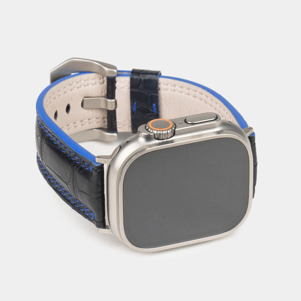 Apple Watch Ultra 49 mm in black alligator leather with blue stitching and edge, with titanium adapters and buckle | 3/4 front view showing the titanium watch case and the square scales of the leather
