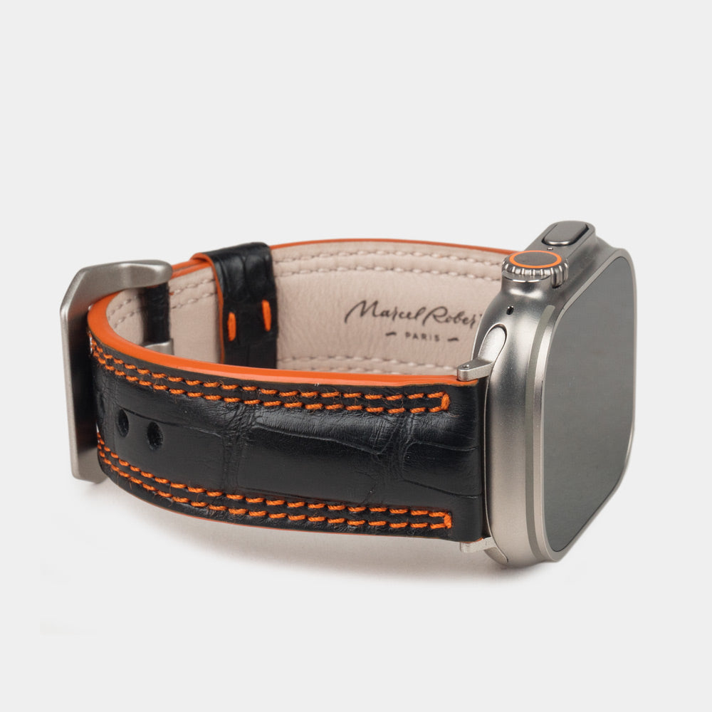 Apple Watch Ultra 49 mm in black alligator leather with orange stitching and edge, with titanium adapters and buckle | 3/4 view showing the leather inner lining and the Marcel Robert signature