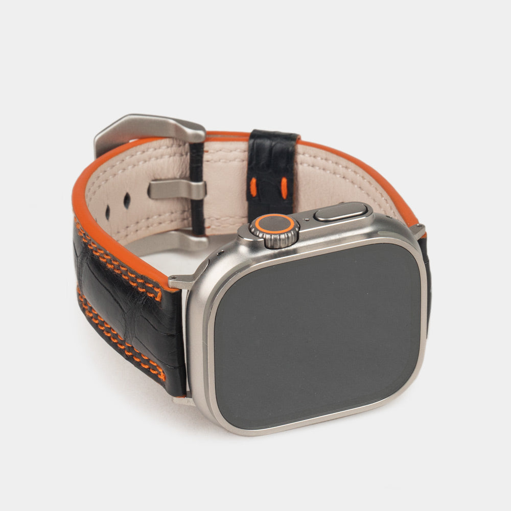 Apple Watch Ultra 49 mm in black alligator leather with orange stitching and edge, with titanium adapters and buckle | 3/4 front view showing the titanium watch case and the square scales of the leather