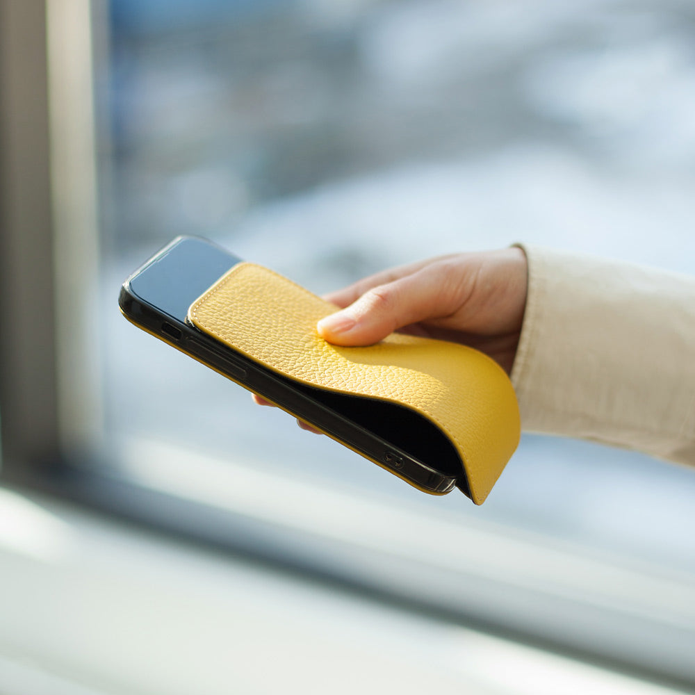 Yellow calfskin leather flip case for iPhone 15 held in one hand by a window, flap folded open behind the phone