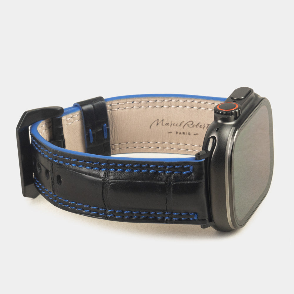 Apple Watch Ultra 49 mm in black alligator leather with blue stitching and edge, with black adapters and buckle | 3/4 view showing the leather inner lining and the Marcel Robert signature