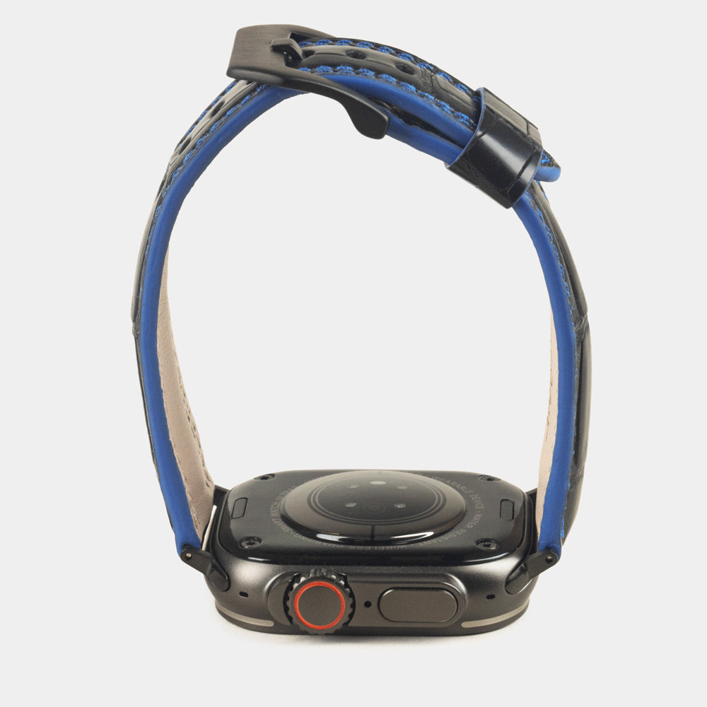 Apple Watch Ultra 49 mm in black alligator leather with blue stitching and edge, with black adapters and buckle | watch case on bottom to see the strap profile and the blue dyed edge